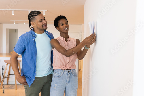 Hanging diverse couple affixing paper sample on white wall near kitchen island at modern home