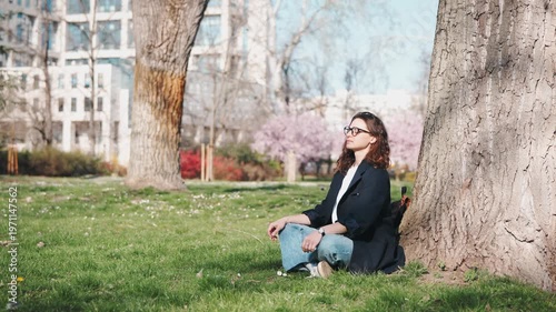 Young business woman in glasses meditating under a tree in a blooming spring park. Calm urban escape, mindfulness, balance, relaxation and mental wellness concept.