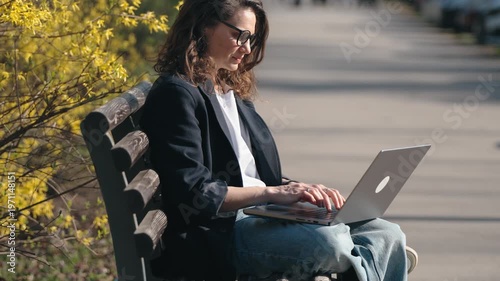 Young woman in glasses working on a laptop while sitting on a bench in a blooming spring park. Remote work, freelance lifestyle, outdoor productivity, modern technology concept.