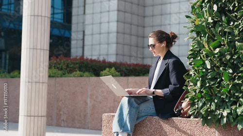 Young business woman having a video conference on laptop outdoors near a modern office building on a warm summer day. Remote work, freelance lifestyle, urban business concept.