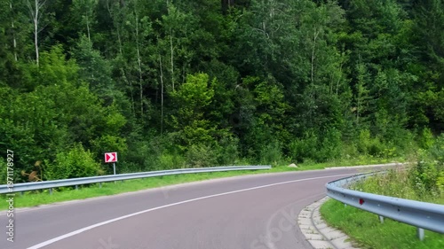 Windshield view forest corner shows chevron signs near silver guardrail lines lane. Car takes tight curve slowing down holding line along inner edge