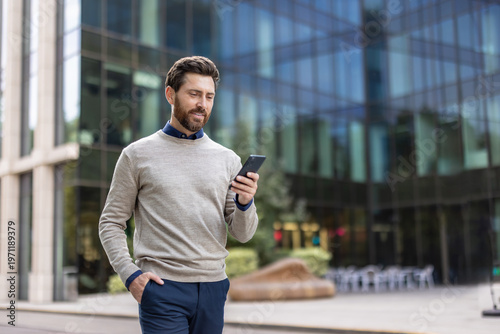 Smiling young man walking near office center and using mobile phone