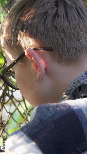 A teenage boy stands behind a metal fence, looking upset and isolated. The scene conveys sadness and bullying at school, highlighting emotional struggle and vulnerability. Vertical video.