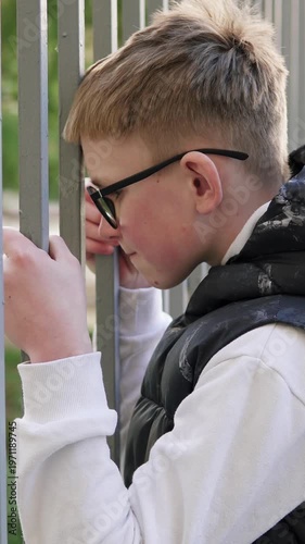 A teenage boy stands behind a metal fence, looking upset and isolated. The scene conveys sadness and bullying at school, highlighting emotional struggle and vulnerability. Vertical video.