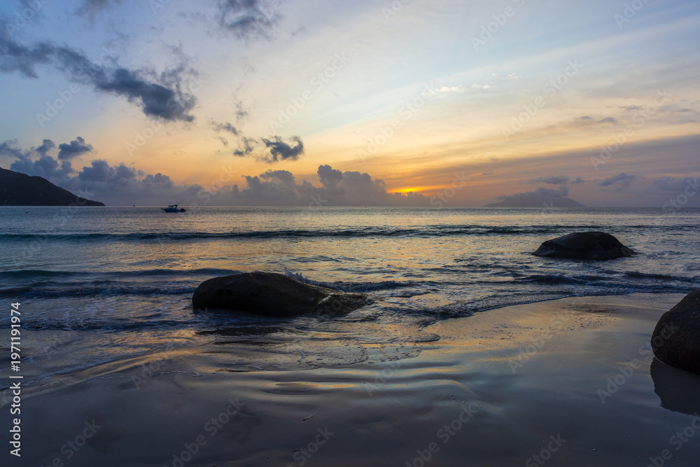 Obraz premium Golden sunset paints pastel skies above a rocky beach as soft waves wash the sand and a lone boat drifts on the calm ocean horizon, creating a peaceful, contemplative seascape mood
