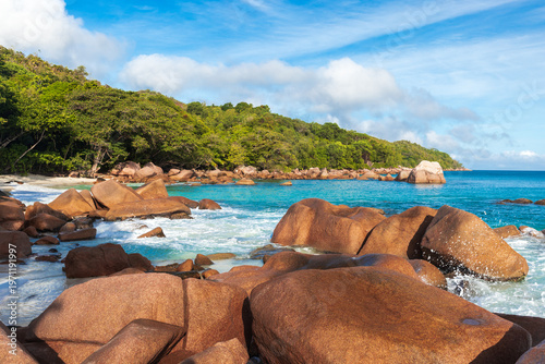 Photography Sunlit tropical beach framed by smooth granite boulders and turquoise water, with waves lapping a lush green shoreline