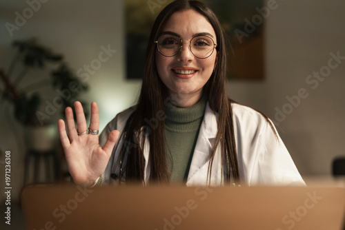 A smiling female doctor in a white coat and glasses waves hello during a video call, offering a friendly greeting.