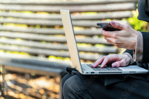 Woman hands typing laptop holding smartphone park bench outdoors