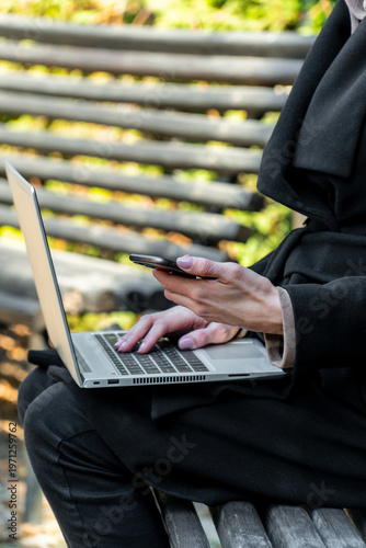 Woman typing on laptop and holding smartphone on park bench