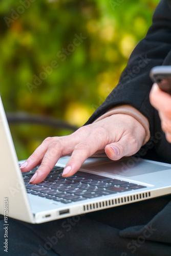 Female hands typing on laptop keyboard outdoors in a park holding smartphone