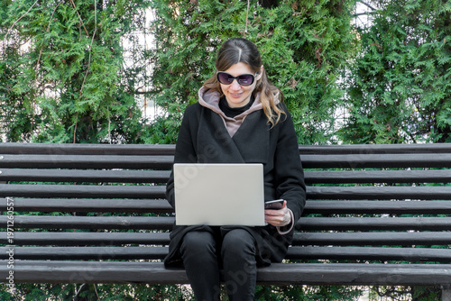 Smiling woman on park bench with laptop and smartphone for remote work freedom