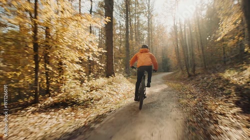 Rear view of a male mountain biker in an orange jacket cycling along a narrow winding trail in a vibrant autumn forest with golden sunlight and lens flare filtering through the trees.