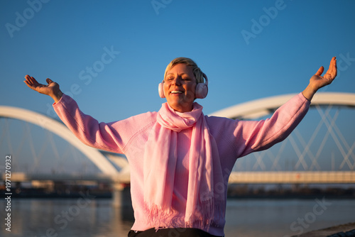 Smiling woman with arms raised enjoying listening to music through wireless headphones while sitting by river in the city on a sunny day.