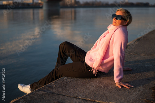 Stylish woman with sunglasses enjoying a peaceful moment by the river in the city, basking in sunlight, feeling free, calm and content after a long day.