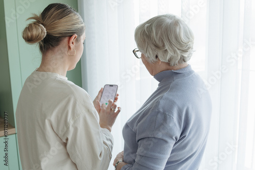 Young woman showing smartphone to senior woman at home near window. Helping elderly person with mobile technology, communication and digital skills, modern lifestyle concept