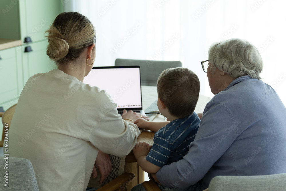 Obraz premium Three generations sitting together at home using laptop. Young caregiver helping senior woman and child with digital technology. Concept of family support, intergenerational learning and modern lifest