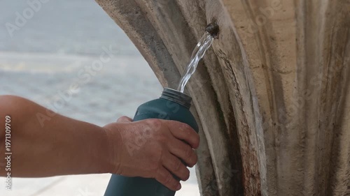 A hand fills a bottle at a drinking fountain while summer dressed tourists stand in the background in warm daylight near the water flow.