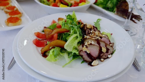 Gourmet duck salad featuring arugula, tomatoes, pear slices, hazelnut, balsamic glaze on white plate in elegant banquet environment