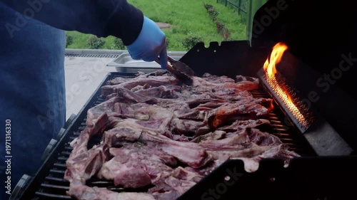 Professional chef wearing blue gloves grilling lamb ribs on sizzling barbecue, manipulating meat with tongs near intense charcoal flames during outdoor cooking process