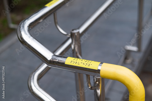 Stainless steel handrail with braille markings and yellow safety coating, accessibility infrastructure outdoors