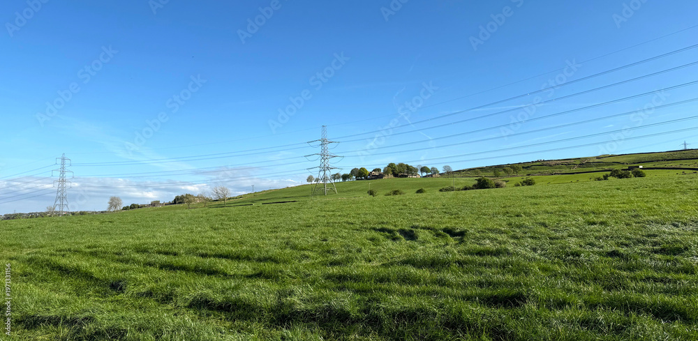 Obraz premium A lush green field stretches under a clear blue sky. Tall pylons tower over the landscape, connecting distant hills near the old village of Thornton, Yorkshire, UK