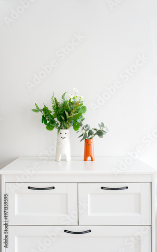Minimalist Scandinavian interior scene with two decorative vases featuring simple smiling faces, filled with green plants and flowers, placed on a clean white cabinet against a neutral wall.