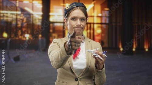 Woman flight attendant pointing finger while holding a toy airplane and wearing beige blazer and red scarf in front of airport terminal with stern expression; authority instruction.