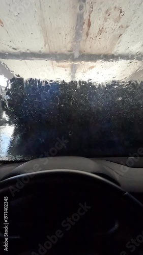 Vertical video of POV from inside a car during automatic wash tunnel, blue foam and water covering the windshield, dashboard visible, brushes and light outside