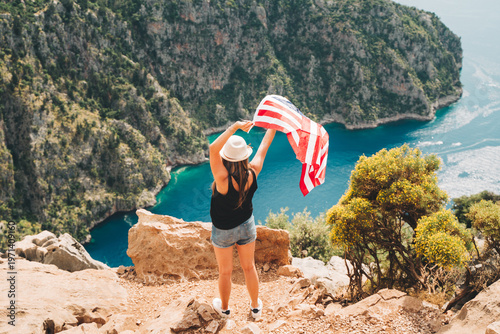 Φωτογραφία Young woman standing on a rock cliff and waving the US flag while looking at sea beneath