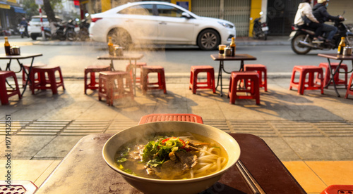 Steaming bowl of traditional Vietnamese beef Pho on a sidewalk cafe table, red plastic stools and city street traffic in the background, authentic street food culture and morning breakfast in Vietnam.