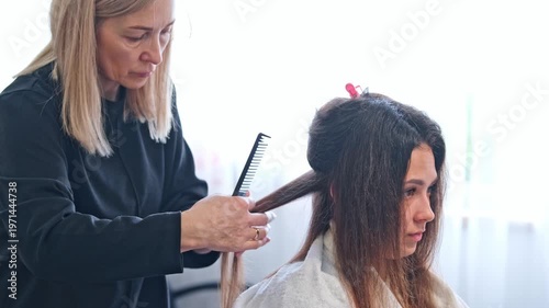 A hair stylist performs keratin hair straightening on a woman's hair using a flat iron. The process takes place in a beauty salon with careful attention to detail.