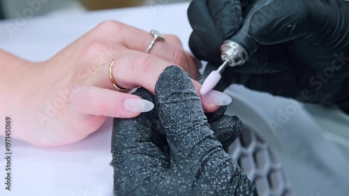 A manicure specialist is using an electric nail drill to file and refine the side of an almond fingernail. This process takes place in a salon setting with focus on nail detail.