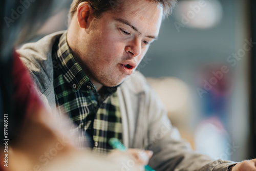 Young student studying intently while writing with a pen at a table in an indoor learning space. A focused student works on a task, showing concentration and engagement in a casual classroom