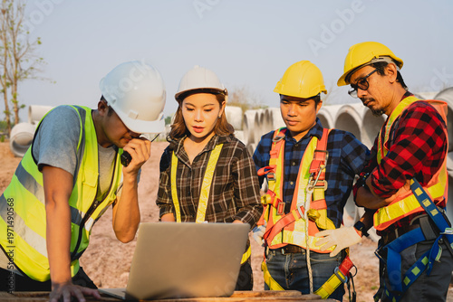 A team of diverse Asian engineers in hard hats and safety vests discussing a project on a laptop at a construction site, featuring concrete pipes and outdoor industrial background.
