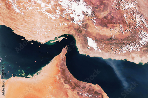 Satellite view of the Strait of Hormuz, connecting the Persian Gulf and Gulf of Oman. High-resolution orbital image featuring arid desert landscapes, deep blue waters, and scattered clouds.