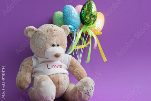 “A plush teddy bear with shiny colorful toy Easter eggs arranged in a bouquet on a lilac background. Easter holidays.”