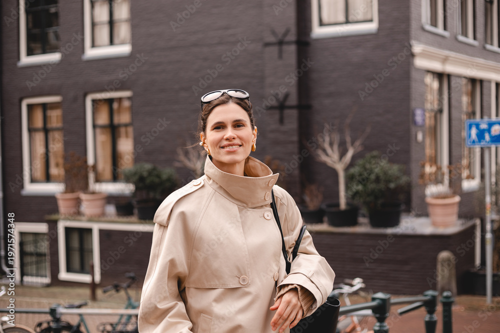 Fototapeta premium Confident and smiling young woman wearing a classic beige trench coat standing on a European city street with historic architecture, bicycles and sunglasses on her head during a cozy autumn day
