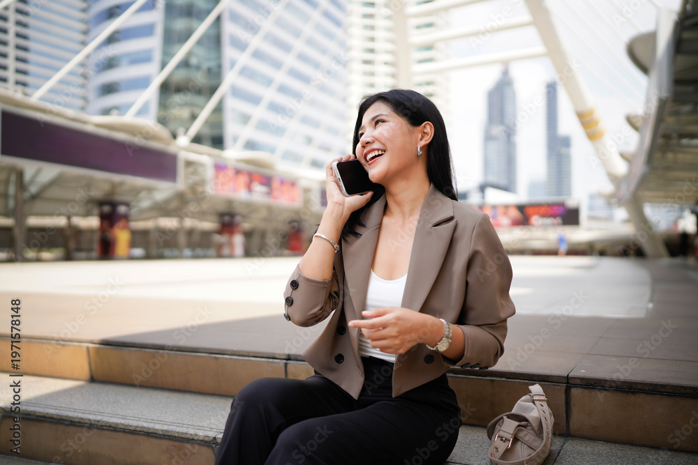 Fototapeta premium Asian businesswoman sitting on stair in downtown talking on mobile phone, skyscrapers cityscape background. Young woman on smartphone happy, woman making business call on cell