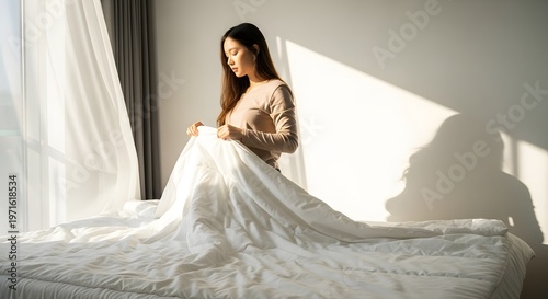 Asian woman tidying white bed in sunny minimalist bedroom. Concept of peaceful morning and home routine.
