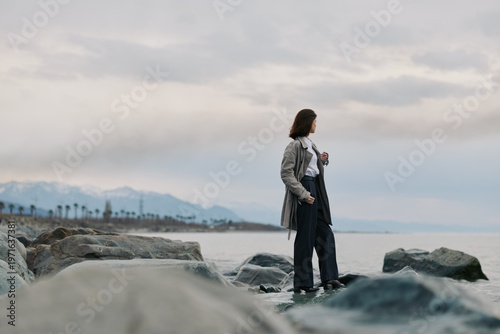 Картината върху платно Woman in casual clothes stands on rocky shore looking at cloudy sky over calm sea