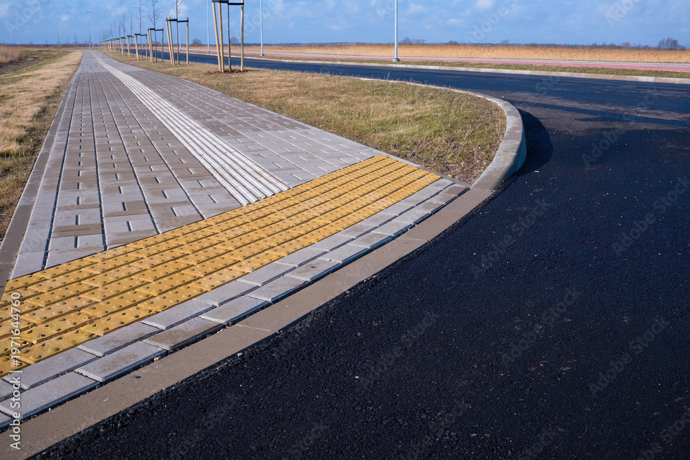 Fototapeta premium Yellow tactile paving for blind and visually impaired people on a new city sidewalk. Modern accessible urban environment with stone tiles and asphalt road. Concept of safety and inclusivity.