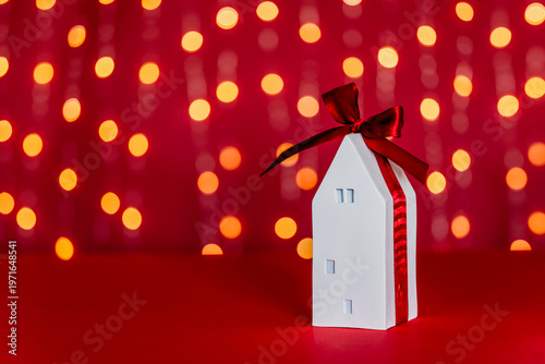 Decorative white house-shaped gift box with red ribbon sits on red surface with blurred warm lights in background