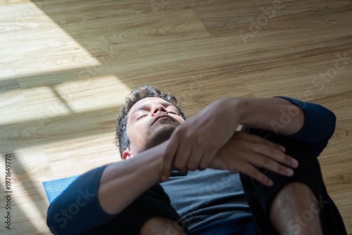 Close-up overhead view of a stressed man lying down and hugging his knees to his chest for rest and introspection in a sunlit room. A dramatic, top-down photograph of a solitary man with dark hair
