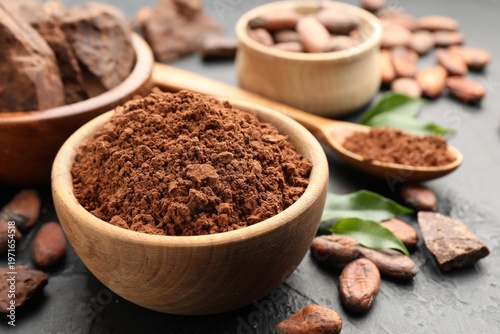 Cocoa powder in bowl, beans and pieces of chocolate on dark textured table, closeup