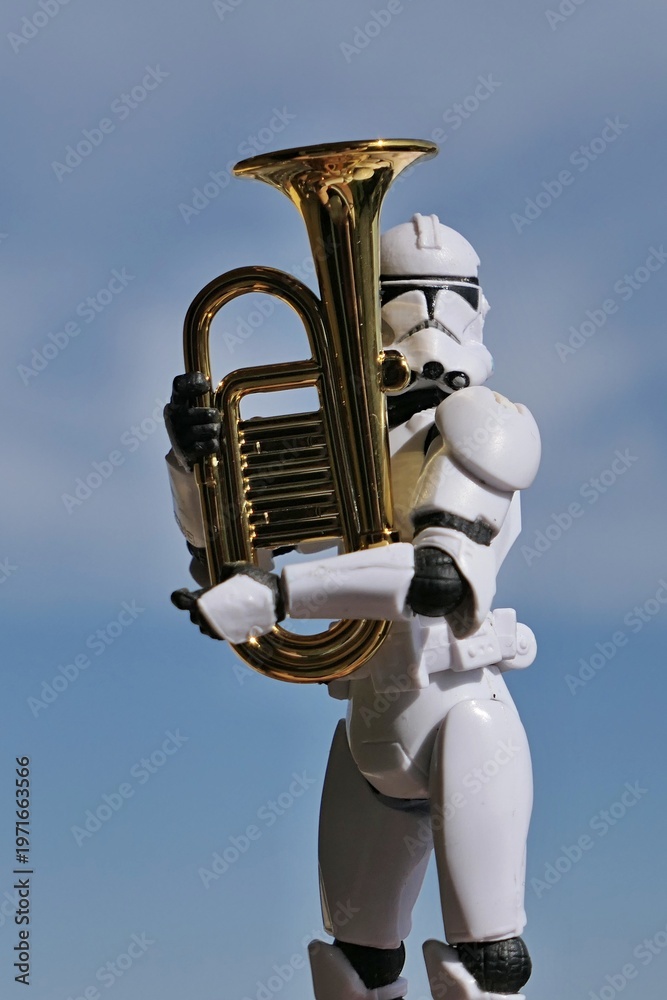 Fototapeta premium Action figure of Star Wars clone trooper in white helmet and armor embracing model of some kind of euphonium brass instrument, blue spring skies with overcast clouds in background. 