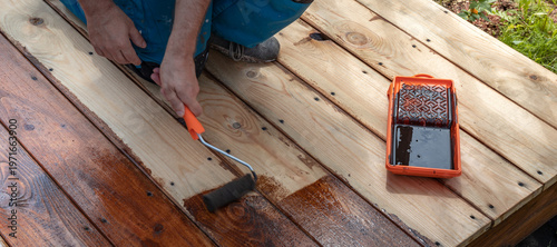 Worker applies wood stain with roller on outdoor deck