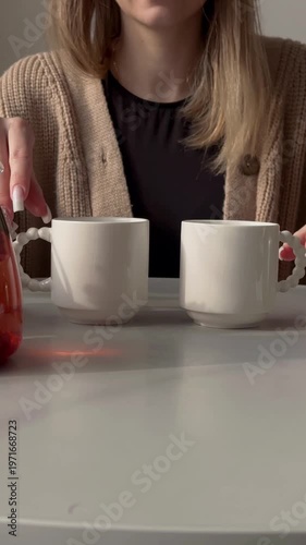 Cozy kitchen white woman pouring tea glass teapot filled with fruit infusion and strawberry slices, ceramic cups on white table,