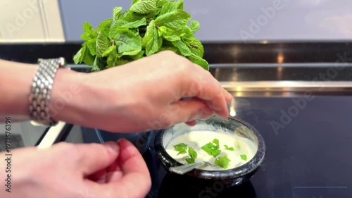 woman putting mint leaves into greek yoghurt in small bowl on kitchen counter