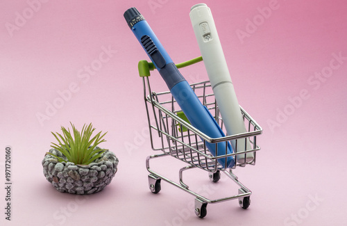 Two medical injection pens, one blue and one white, placed inside a miniature metal shopping cart on a pink background, symbolizing the high cost of healthcare and diabetes management