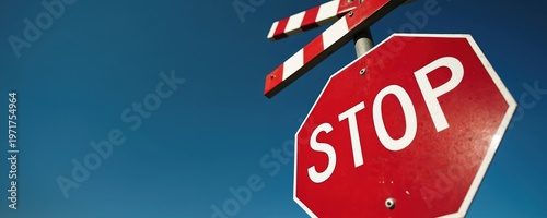 Red stop sign with striped barrier at railway crossing against clear blue sky. Traffic control symbol for train tracks, caution, and safety.